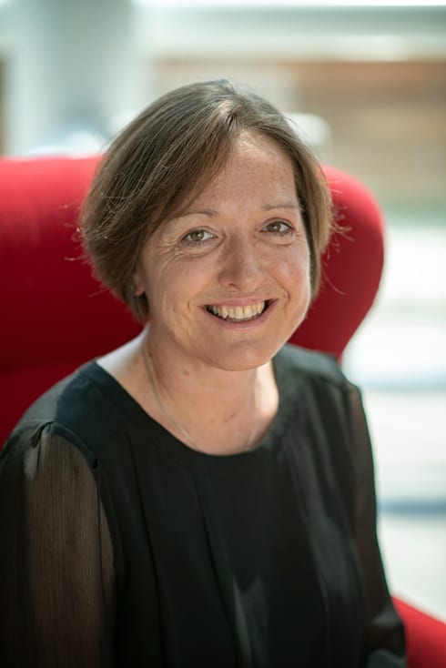 Colour photograph of Gilly Body - a white woman with dark hair, wearing a black top, sitting on a red chair and smiling towards the camera.